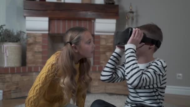 Słodki biały chłopiec w okularach VR rozmawia z ładną dziewczyną i rozgląda się. Brat i siostra bawią się nowym urządzeniem w domu. Weekendy, zajęcia rekreacyjne, odpoczynek, dzieciństwo. — Wideo stockowe