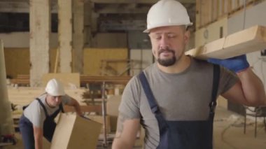 Beyaz koruyucu miğferli kaslı beyaz kaslı bir adam inşaat alanında tahta bir kalasla yürüyor. Arkasındaki ağrıdan dolayı inşaatçı durdu, meslektaşı yardım etmek için yukarı koştu..