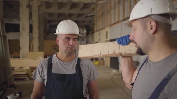Deux constructeurs caucasiens adultes portant des casques blancs protecteurs parlent sur le chantier de construction. Un ouvrier tournant avec une planche, son collègue esquivant . — Video