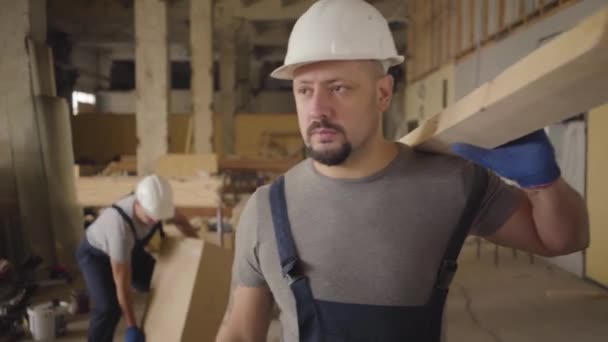 Homme caucasien fort en casque blanc et uniforme bleu marchant avec une planche de bois sur le chantier de construction et agitant la main. Builder se sent mal et s'arrête, son collègue se précipite pour aider . — Video