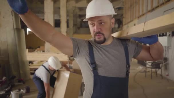 Homme caucasien adulte au casque blanc et uniforme bleu marchant sur le chantier avec une planche de bois. Builder arrêter en raison de douleurs aiguës dans le dos, un autre travailleur se précipite pour aider . — Video