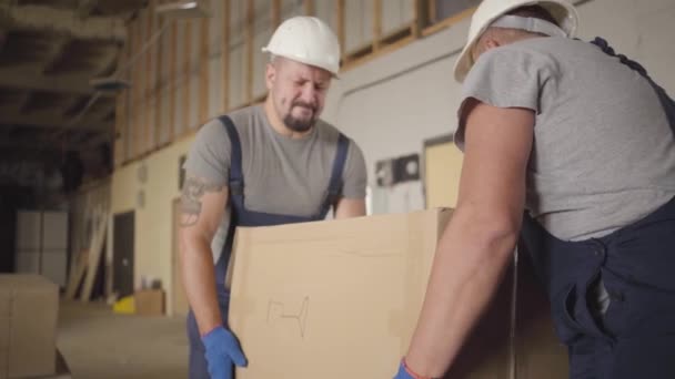 Dois fortes trabalhadores caucasianos musculares pegando caixa de papelão pesado e carregando-o no local de construção. Construtores masculinos adultos em capacetes de proteção e uniforme trabalhando dentro de casa. Trabalho duro . — Vídeo de Stock