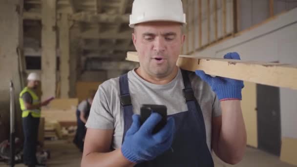 Close-up portret van vrolijke blanke man in beschermende helm kijkend naar zijn smartphone en lachend. Gespierde man op de bouwplaats met houten plank. Volwassen mannelijke bouwer aan het werk. — Stockvideo