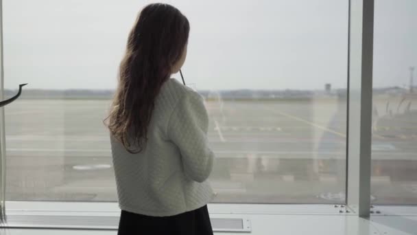 Achteraanzicht van een blank meisje met lang krullend haar kijkend naar het grote raam van de luchthaven en het drinken van sap. Kind wachtend op het vertrek of op familie die komt. Reizen, toerisme, weekenden. — Stockvideo