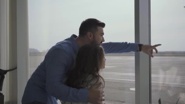 Πλευρική άποψη του νεαρού γενειοφόρου Καυκάσου που δείχνει αεροπλάνα από το παράθυρο του αεροδρομίου. Μικρό όμορφο κορίτσι περιμένει την αναχώρηση ή την άφιξη με τον πατέρα. Ταξίδια, τουρισμός, Σαββατοκύριακα. — Αρχείο Βίντεο