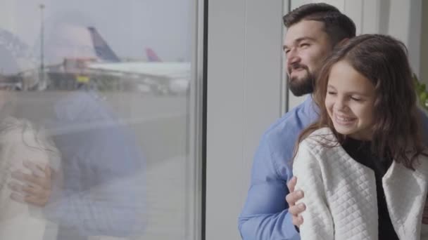 Közelkép egy boldog fehér férfiról, aki a lányával ül a reptér ablakpárkányán, és a kifutópályát nézi. Egy csinos kislány beszél apával. Utazás együtt, turizmus, utazás. — Stock videók