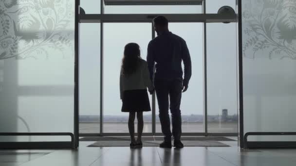 Back view silhouettes of adult man and little girl standing at the airport door and looking at the runway. Father and daughter waiting for the airplane takeoff. Travelling together, tourism, journey. — Stock Video