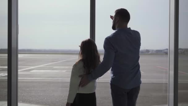 Pohled zezadu na bělochy a dívčí siluety, jak se dívají ze skleněných letištních dveří na přistávací dráhu. Otec a dcera se drží za ruce na čele. Rodina čeká na vzlet letadla. — Stock video