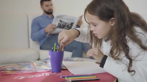 Retrato de linda chica caucásica dibujando en la sala de espera VIP como su padre leyendo el periódico en el fondo. Joven mirando a su hija y riéndose con ella. Esperando la salida del avión . — Vídeo de stock