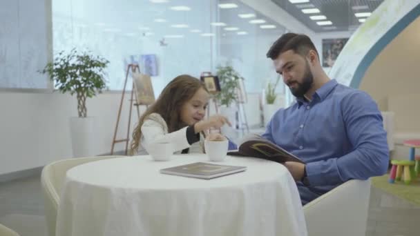 Богатый кавказский мужчина и маленькая девочка сидят в зале ожидания аэропорта VIP, смотрят на фотографии и пьют чай или кофе. Отец и дочь из бизнес класса отдыхают перед отъездом . — стоковое видео