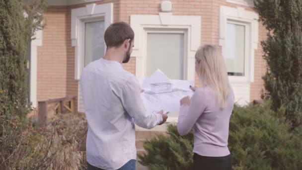 Maschio architetto caucasico e la sua assistente femminile esaminando cianografie di fronte alla casa e dando il cinque. Appaltatore professionista insegnare nuovi segreti dipendenti del lavoro . — Video Stock