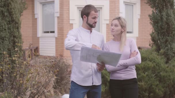 Knappe blanke professionele ontwerper die met zijn assistent voor het huis staat en blauwdrukken onderzoekt. Beroepsopbouwer geeft les aan nieuwe werknemer. — Stockvideo