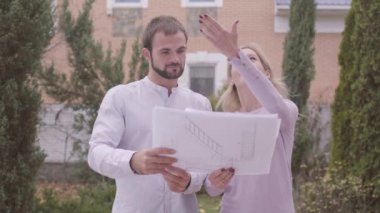 Genç, beyaz bir kadın müşterisine mimari çizimler ve çalışmaların sonuçlarını gösteriyor. Profesyonel tasarımcı, memnun müşteriyle birlikte evin önünde duruyor. İnsanlar beşlik çakıyor..