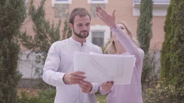 Young attractive Caucasian woman showing her client architectural drawings and result of work. Professional designer standing with satisfied customer in front of the house. People giving high five. — Stock Video