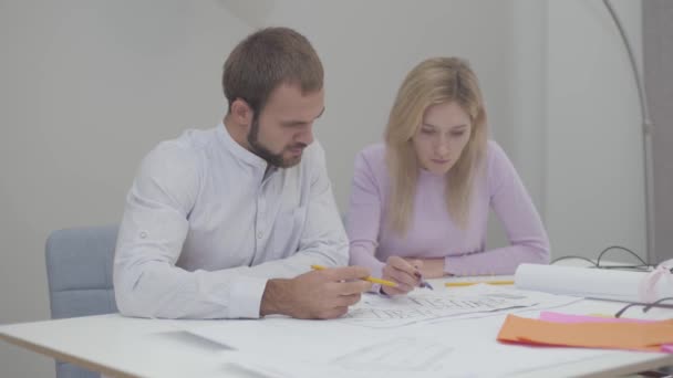 Professionele blanke mannelijke ontwerper doceert nieuwe werknemer in het kantoor. Jonge mooie vrouw tekent blauwdrukken. Specialisten die ideeën op papier uitvoeren. — Stockvideo