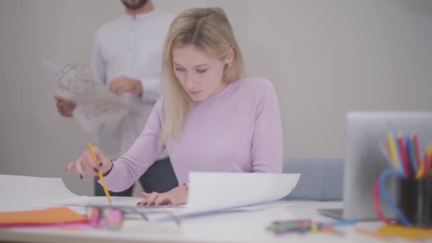 Portrait of beautiful blond Caucasian woman drawing blueprints at the office. Her male colleague standing at the background with drafts. Professional architect showing his works to coworker. — Stockvideo