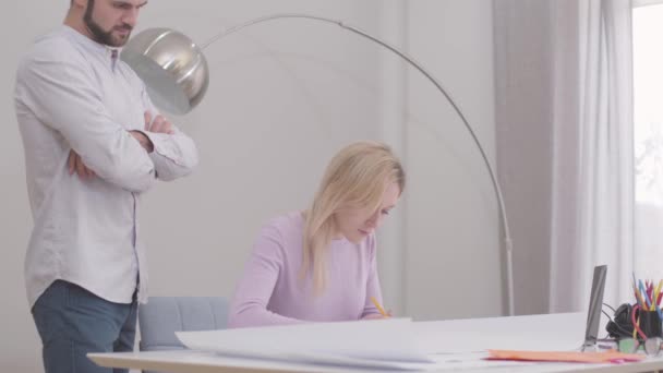 Vista lateral de la joven arquitecta caucásica dibujando un plano en la oficina. Su colega masculino controlando el proceso. Diseñadores profesionales trabajando en su proyecto. Especialistas en el lugar de trabajo . — Vídeo de stock