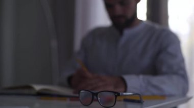 Arka planda evraklarla çalışan bulanık bir beyaz adam. Erkek profesyonel mimar masada oturarak proje yapıyor. Ön planda duran zarif siyah gözlüklere odaklanmış..