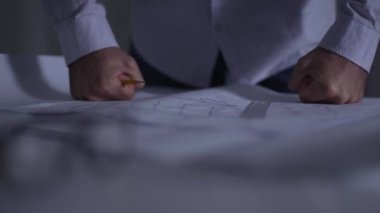 Masada yatan mimari çizimlerin üzerine öfkeyle dikilmiş beyaz erkek yumruklarının yakın çekimi. Yetişkin mimar işinden memnun olmayan kalemleri atıyor. Projenin uygulanmasında sorunlar.