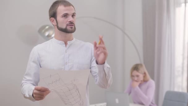 Jonge zelfverzekerde blanke man die in de kamer rondkijkt en aantekeningen maakt in zijn ontwerpen. Professionele architect creëert het idee van interieurontwerp. Zijn collega tekent blauwdrukken op de achtergrond. — Stockvideo