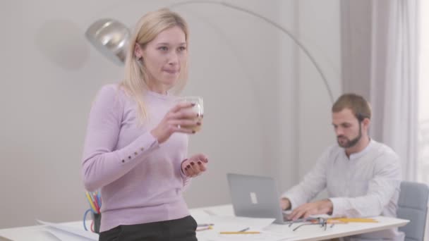 Mulher branca bonita bebendo cappuccino ou latte, voltando e conversando com seu colega masculino. Homem barbudo fechando laptop e sorrindo. Arquitetos profissionais que têm pausa . — Vídeo de Stock