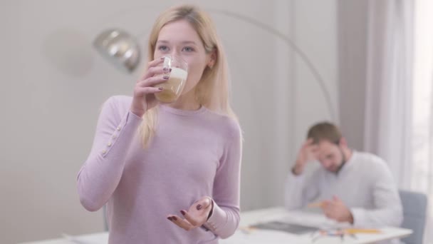 Egy fiatal szőke nő portréja, aki cappuccinót iszik vagy lattét és mosolyog. Professzionális építészek szünetelnek. A férfi kollégája tervrajzokat rajzolt a háttérben.. — Stock videók