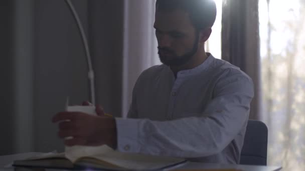 Hombre caucásico guapo sentado en el interior a la luz del sol de la noche y tomando notas en el cuaderno. Arquitecto profesional masculino haciendo proyecto sentado en la mesa . — Vídeos de Stock