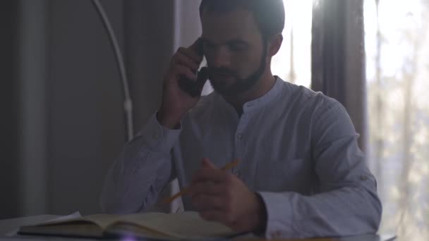 Jeune homme caucasien parlant sur un téléphone intelligent dans la lumière du soleil du soir et de prendre des notes dans un carnet. Architecte professionnel masculin faisant projet assis à la maison ou au bureau . — Video