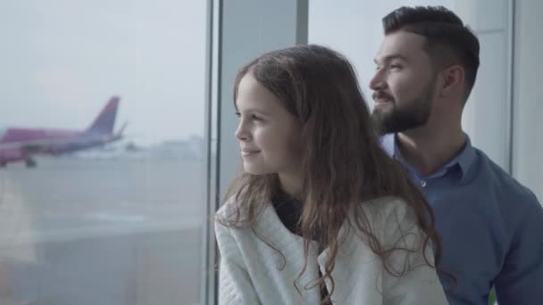 Close-up faces of pretty Caucasian girl with curly hair and adult bearded man looking at airplane takeoff. Father and daughter sitting at the airport windowsill and looking at the runway. Tourism. — Stock Video