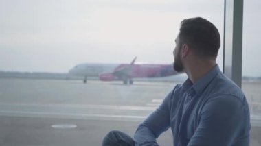 Uçağın kalkışına bakan yakışıklı sakallı adamın yan görüntüsü. Genç beyaz adam havaalanının pencere eşiğinde oturuyor ve pencereden podyuma bakıyor. Seyahat, turizm.
