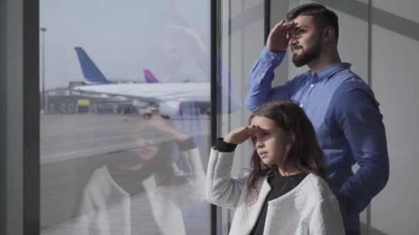 Vista lateral de primer plano del hombre caucásico y la niña mirando por la ventana del aeropuerto de vidrio en la pista. Padre e hija tomados de la mano en la frente. Familia esperando el despegue del avión . — Vídeos de Stock