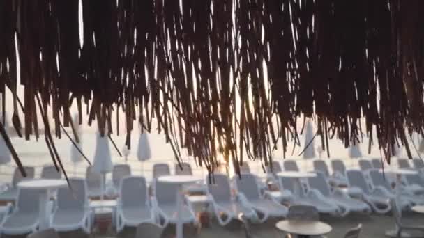 Promienie słoneczne świecące przez słomkowy parasol na wschodniej plaży. Słoneczny brzeg piasku z białymi leżakami w tle. Spokojny kurort w Turcji. — Wideo stockowe