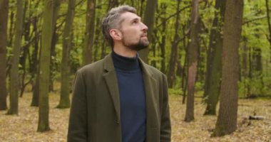 Gri saçlı beyaz bir adamın ormanda dikilip yukarı baktığı yakın plan. Sakin bir sonbahar gününün tadını çıkaran sıradan giysili bir adam. Sinema 4k görüntüleri Prores Hq.