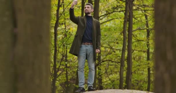 Sonriente hombre de pelo gris caucásico de pie entre los árboles en el bosque de otoño y tomando fotos en su teléfono inteligente. Guapo turista pasar vacaciones al aire libre. Cinema 4k metraje ProRes HQ . — Vídeos de Stock