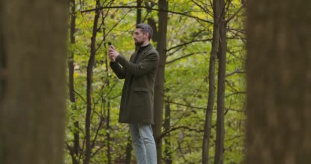 Hombre caucásico sonriente con el pelo gris de pie entre los árboles en el bosque de otoño y tomar fotos en su teléfono inteligente. Guapo turista pasar vacaciones al aire libre. Cinema 4k metraje ProRes HQ . — Vídeos de Stock