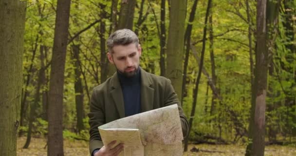 Красивый кавказский турист, стоящий в осеннем лесу между деревьями и смотрящий на карту. Уверенный в себе человек ищет лучший способ уйти. Съемки в кинотеатре 4k . — стоковое видео
