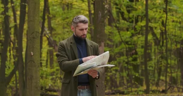 Elveszett ősz hajú fehér férfi áll az őszi erdőben, nézi a térképet és rázza a fejét. Zavart turista keresi a legjobb utat. Mozi 4k felvételek Prores Hq. — Stock videók