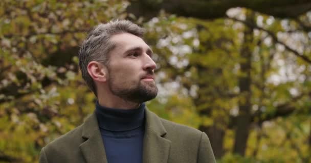 Portrait of handsome Caucasian man with gray hair and brown eyes standing in forest and looking around. Confident tourist in casual clothes enjoying autumn day outdoors. Cinema 4k footage ProRes HQ. — Stock Video