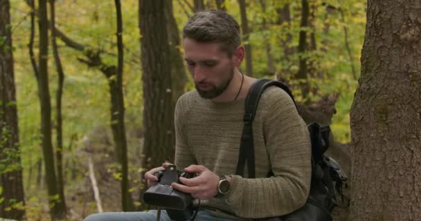 Красивый взрослый кавказский турист сидит в осеннем лесу и проверяет свою камеру. Уверенный седовласый турист проводит выходные на открытом воздухе. Съемки в кинотеатре 4k . — стоковое видео