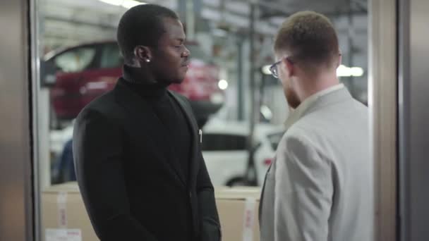 Retrato de jovem afro-americano bonito conversando com revendedor de carros no showroom. Cara de sucesso em roupas pretas discutindo compra de carro com vendedor caucasiano. Concessionária de automóveis, empresa de automóveis . — Vídeo de Stock