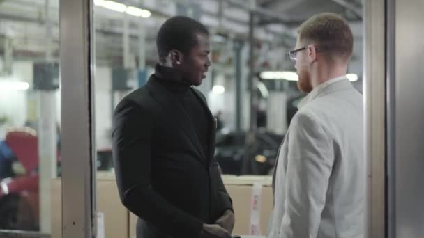 Porträtt av framgångsrika manliga afroamerikanska bilhandlare som ger nycklar till byuern. Ung självsäker vit man i kostym köper ny bil i showroom. Bilhandel, bilindustri. — Stockvideo