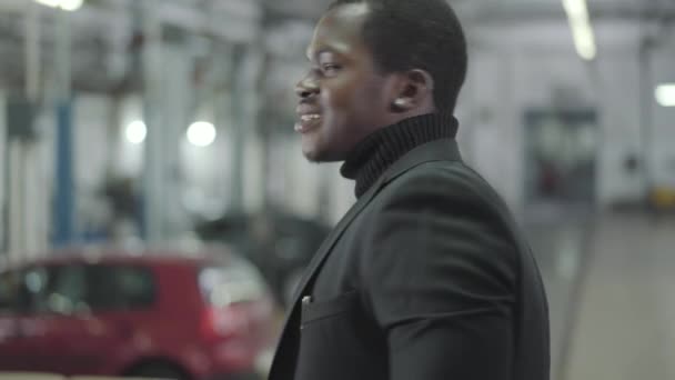 Mladý usměvavý Afroameričan se otáčí ke kameře a mluví na mobilu v autosalonu. Sebevědomý prodejce aut na jeho pracovišti. Obchod s auty, obchod s auty. — Stock video