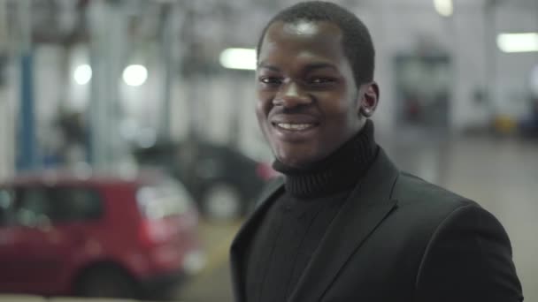 Bonito afro-americano com olhos castanhos a virar-se para a câmara e a sorrir. Traficante de carros masculino confiante ficar em seu local de trabalho. Concessionária de automóveis, empresa de automóveis . — Vídeo de Stock