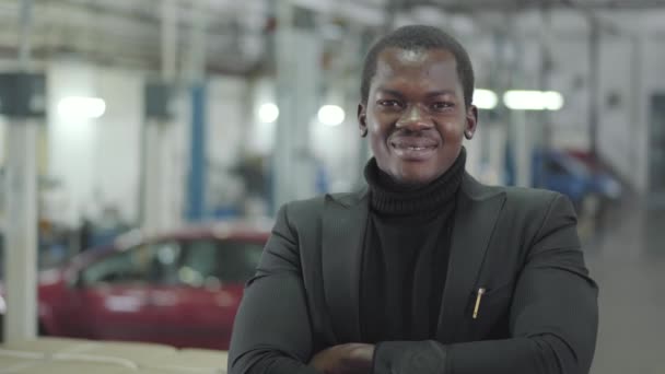 Молодий успішний афроамериканський бізнесмен дивиться на фотоапарат і посміхається. Власник автомобільної виставкової зали стоїть на задньому плані нових автомобілів. Автомобільний дилер, автомобільний бізнес. — стокове відео