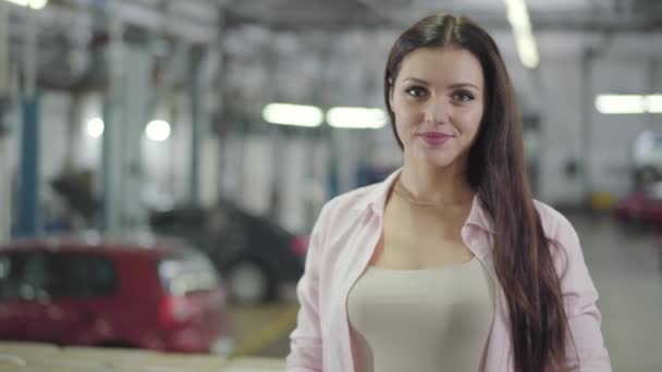 Retrato de confianza concesionario de coches caucásicos mirando a la cámara y sonriendo. Joven mujer exitosa de pie en la sala de exposición de automóviles. Hermosa chica satisfecha con su trabajo. concesionario de coches, negocio de automóviles . — Vídeos de Stock