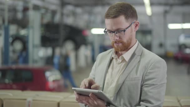 Portret rudego białego mężczyzny w okularach, handlującego online za pomocą tabletu, patrzącego w kamerę i gestykulującego "tak" trzęsąc głową. Salon samochodowy, biznes samochodowy. — Wideo stockowe