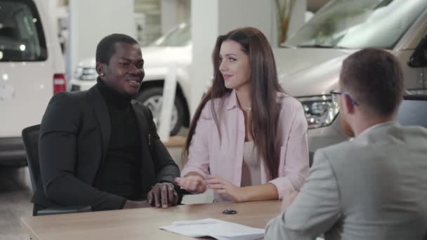 Joven hermosa mujer caucásica hablando con concesionario de coches y sosteniendo maridos mano. Familia Biracial comprando un automóvil nuevo. Feliz pareja multirracial haciendo trato en la sala de exposición. concesionario de coches, negocio de automóviles . — Vídeos de Stock