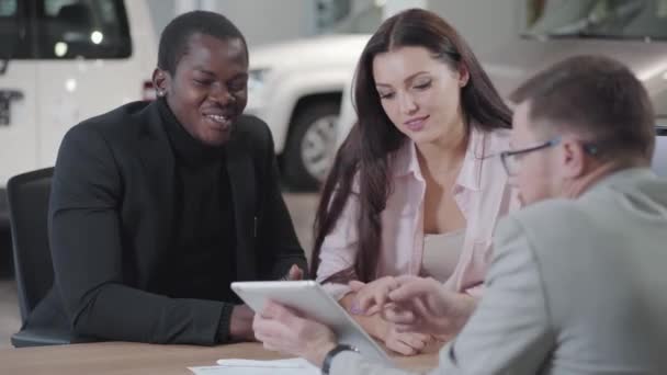 Man Kaukasische auto dealer met rode baard in bril uit te leggen deal details aan jonge multiraciale paar. Succesvolle Afro-Amerikaanse zakenman koopt nieuwe auto voor zijn vriendin of vrouw. — Stockvideo