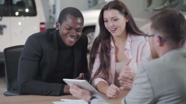 Vista posterior del joven concesionario de automóviles caucásicos mostrando automóviles en su tableta a la joven pareja multirracial. Exitoso hombre de negocios afroamericano que compra un coche nuevo para su novia o esposa en la sala de exposición . — Vídeos de Stock