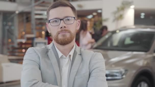 Närbild av unga kaukasiska bilhandlare gestikulerar ja genom att skaka huvudet och le. Självsäker framgångsrik säljare poserar i bakgrunden av biracial par beundrar sin nya bil. — Stockvideo